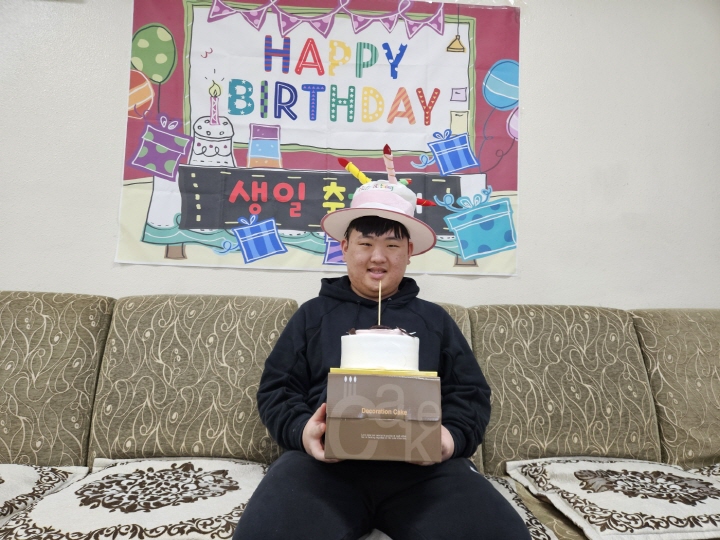 생일을 맞은 주간보호센터 이용인이 생일축하 현수막 앞에 앉아 생일을 기념하는 모자를 쓰고 케익을 든 채로 웃고 있는 모습입니다. 