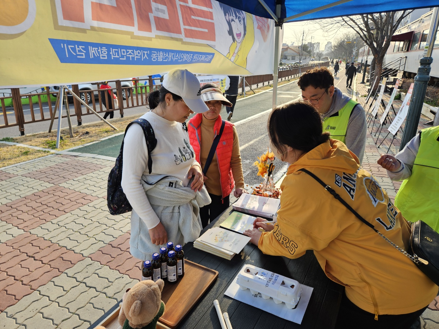 홍보부스 안에서 설명하고 있는 직원과 집중해서 듣고 있는 지역주민의 모습이 담긴 사진입니다. 