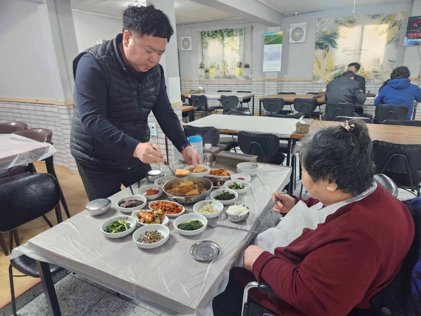 어머님과 함께 식당에서 식사를 하며 앞접시에 국을 담아 드리는 사회복지사와 앞자리에 앉아 그 모습을 보고 있는 어머님의 모습입니다. 