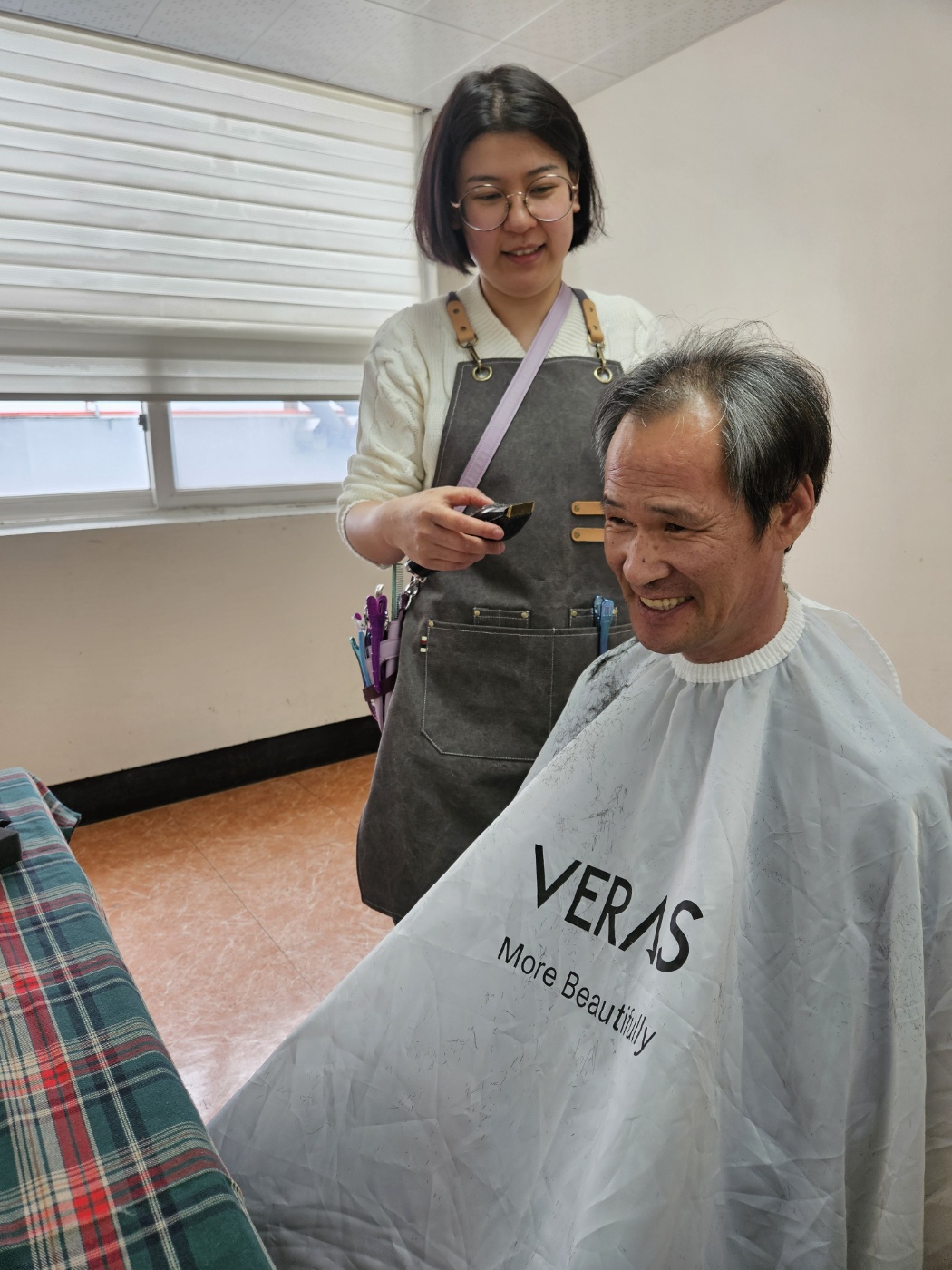 미용도구인 바리깡을 들고 있는 봉사자와 밝게 웃으며 거울을 보고 있는 남성이용인의 모습입니다. 