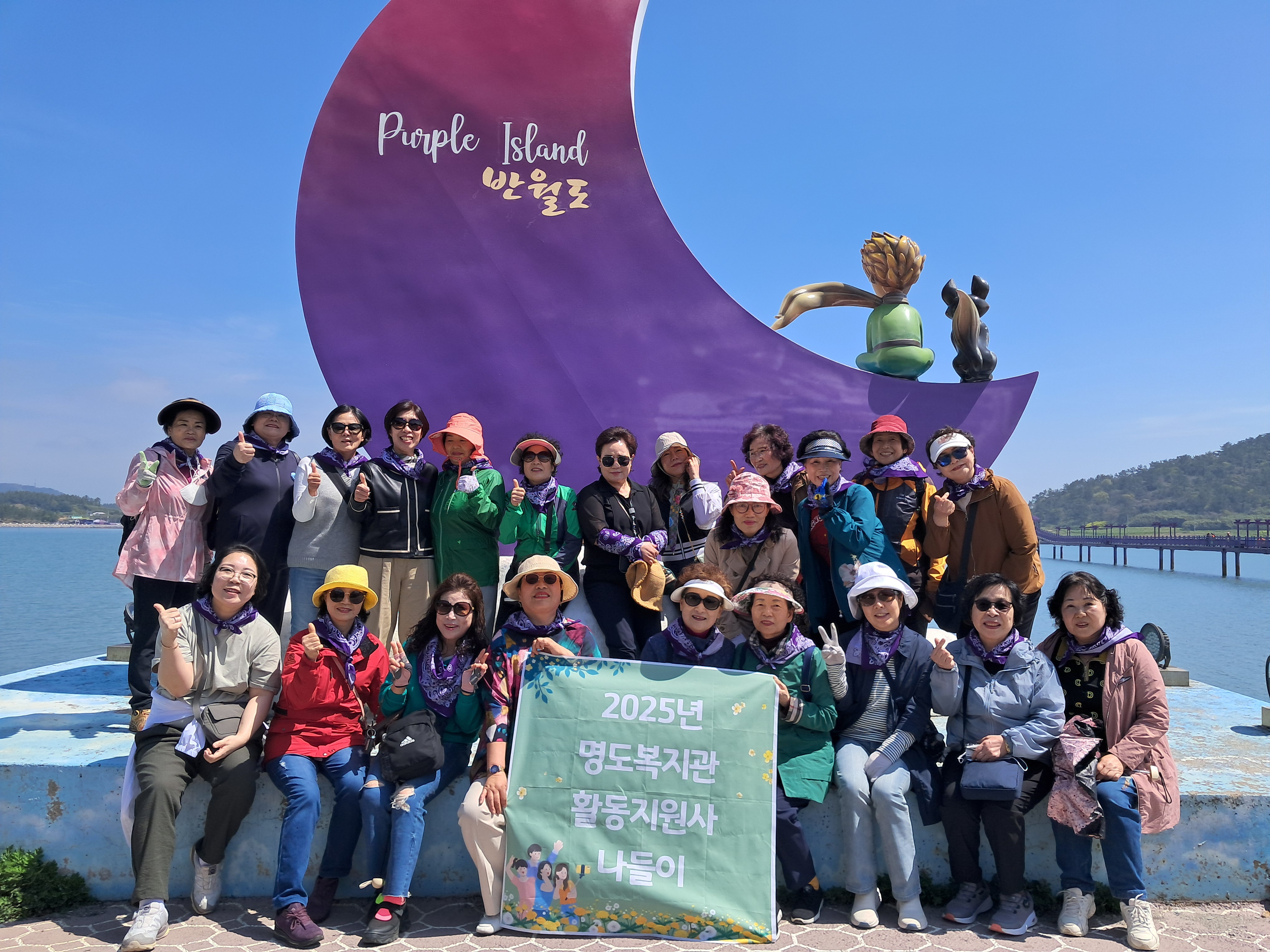 반월도를 나타내고 있는 조형물앞에서 2025년 명도복지관 활동지원사 나들이 현수막을 펴고 함께 찍은 단체사진입니다. 