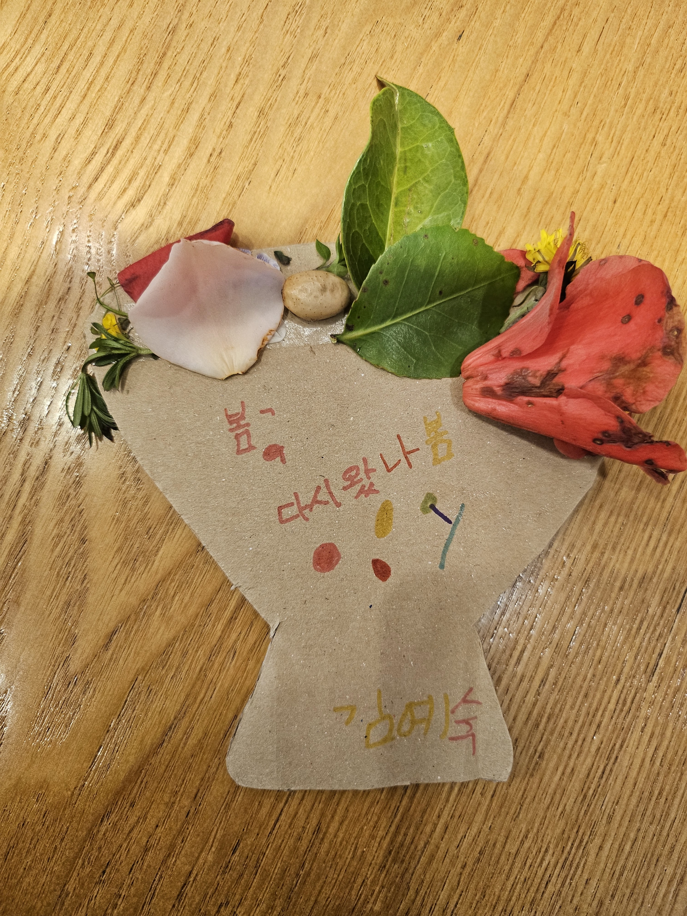 떨어진 꽃잎과 나뭇잎으로 종이화병을 꾸민 작품 사진입니다. 종이화병에는 봄, 다시 왔나봄. 김예숙이라고 적혀있습니다. 