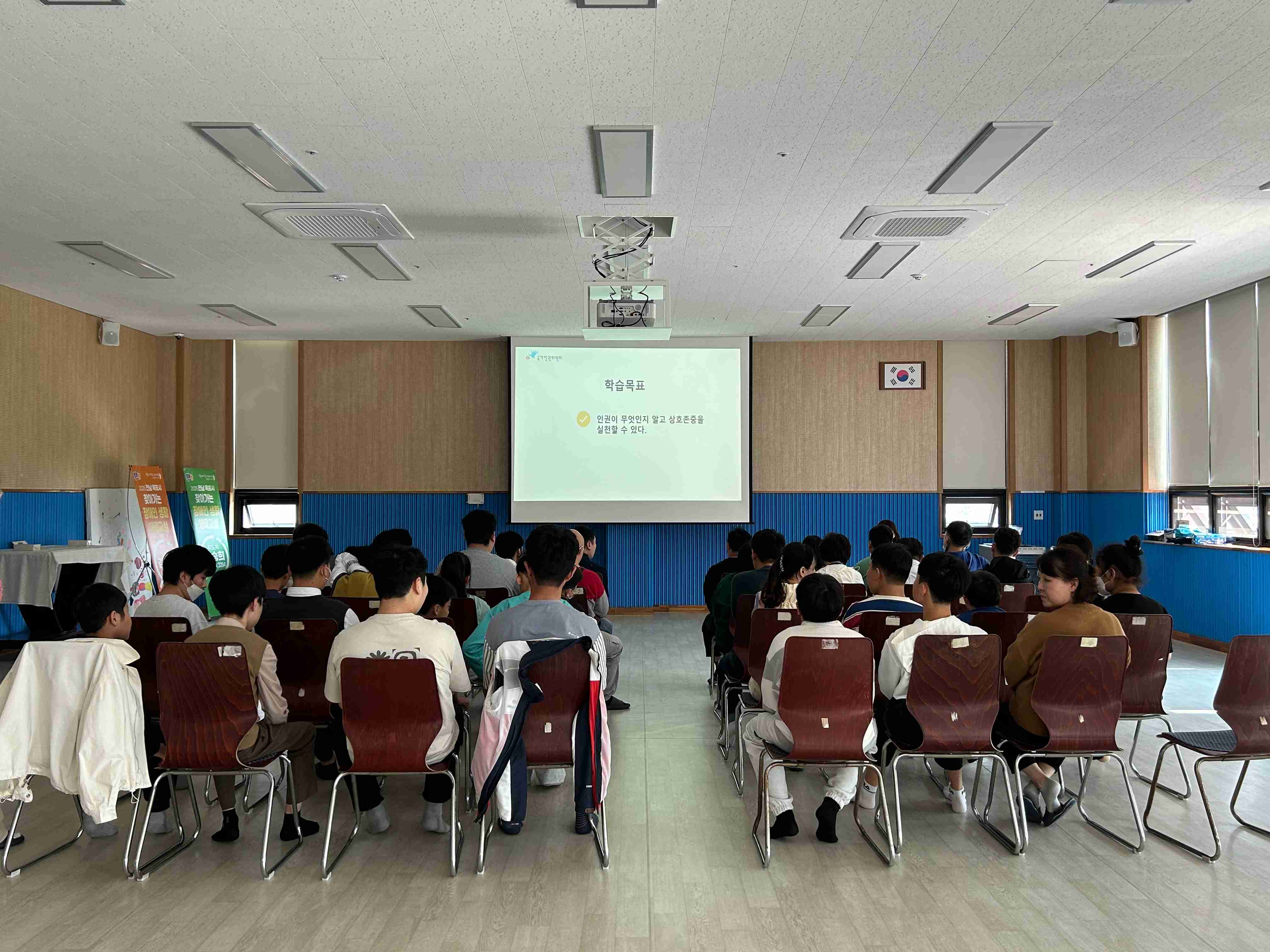 4동 강당에서 많은 이용인들이 의자에 앉아 강의를 듣고 있는 뒷모습입니다. 