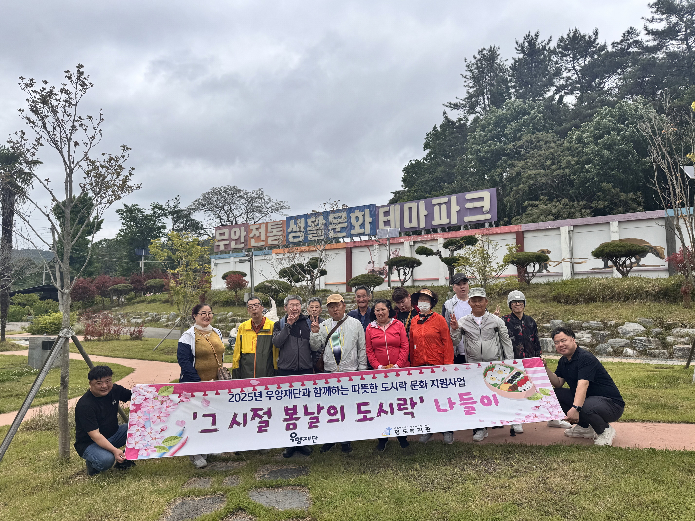 2025년 우양재단과 함께하는 따뜻한 도시락 문화 지원사업. '그 시절 봄날의 도시락' 나들이 현수막을 들고 담당 사회복지사들과 참여자들이 무안전통생활문화테마파크앞에서 함께 찍은 단체사진입니다. 