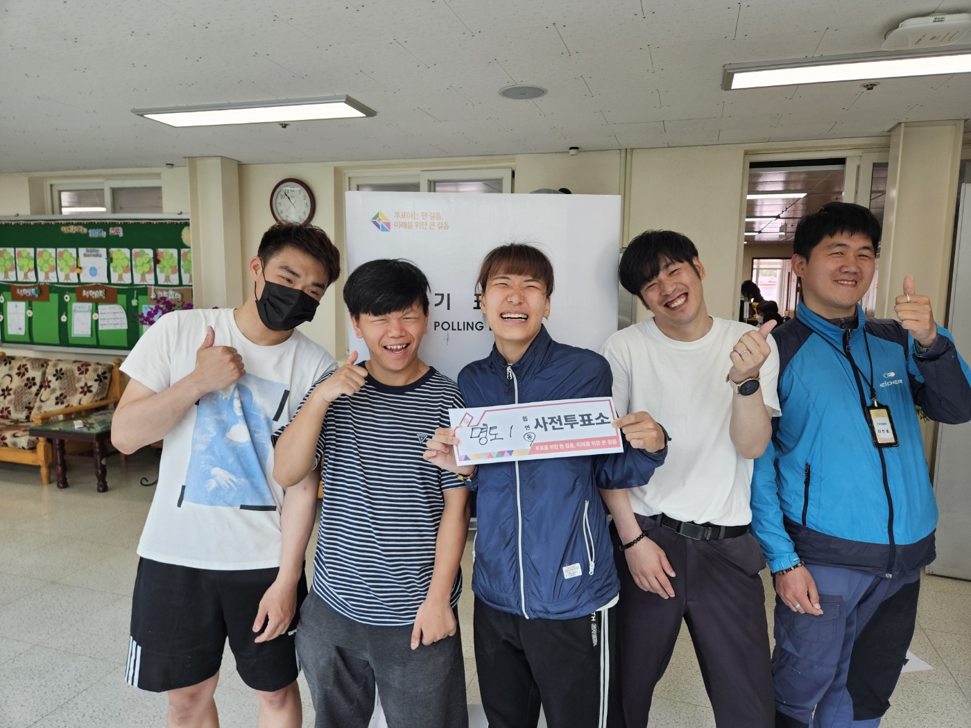 기표소를 배경으로 명도1동 사전투표소라는 안내판을 들고 활짝 웃으며 기념사진을 찍은 이용인 5명의 모습입니다. 