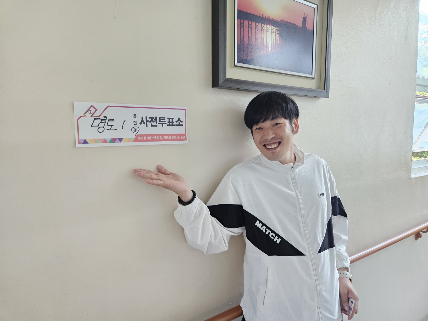명도1동 사전투표소라고 적힌 안내판을 가리키며 활짝 웃는 표정으로 촬영한 이용인의 사진입니다. 