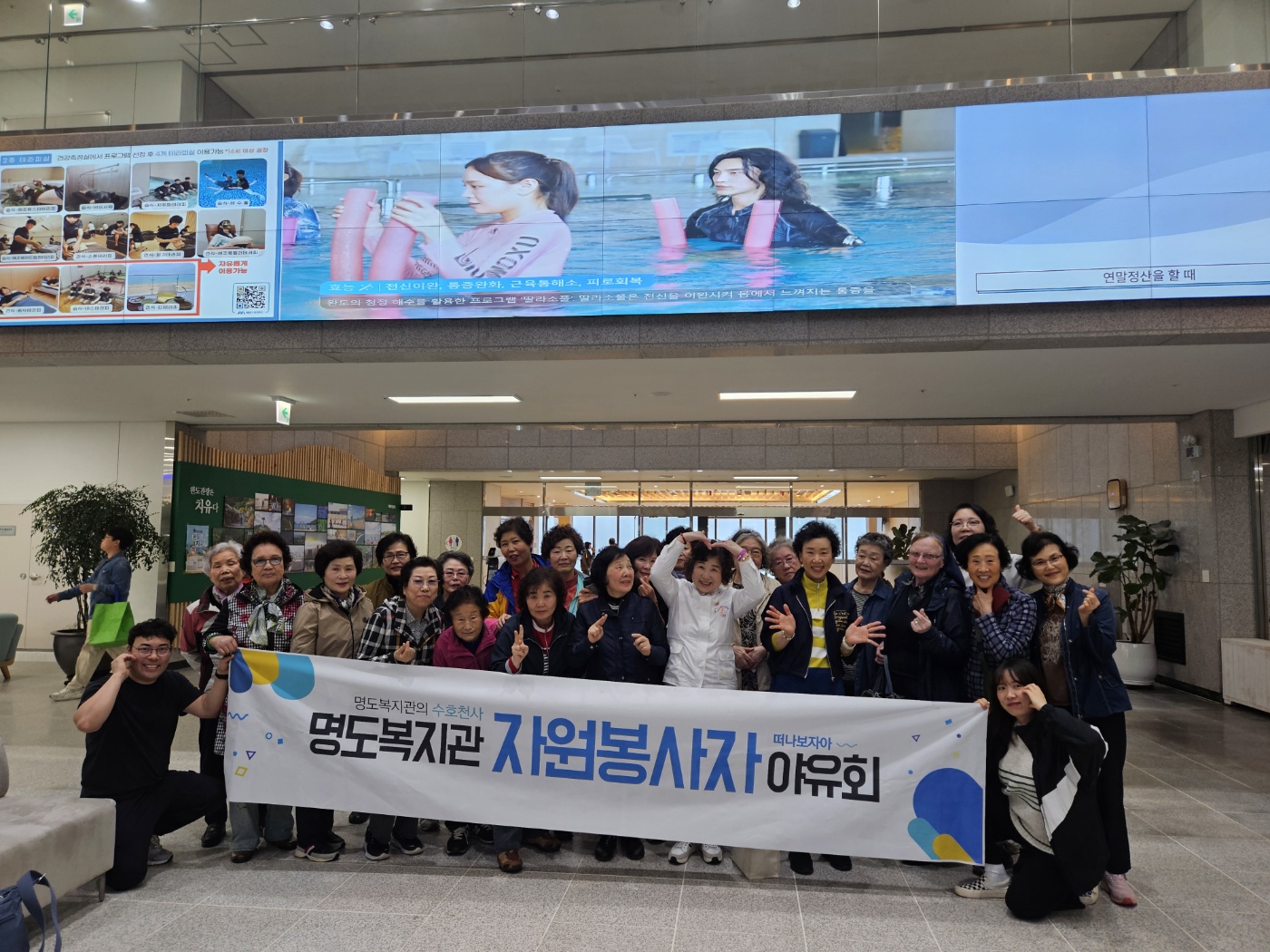 해양치유센터 로비에서 '명도복지관의 수호천사, 명도복지관 자원봉사자 야유회'라고 적힌 현수막을 들고 찍은 단체사진입니다. 