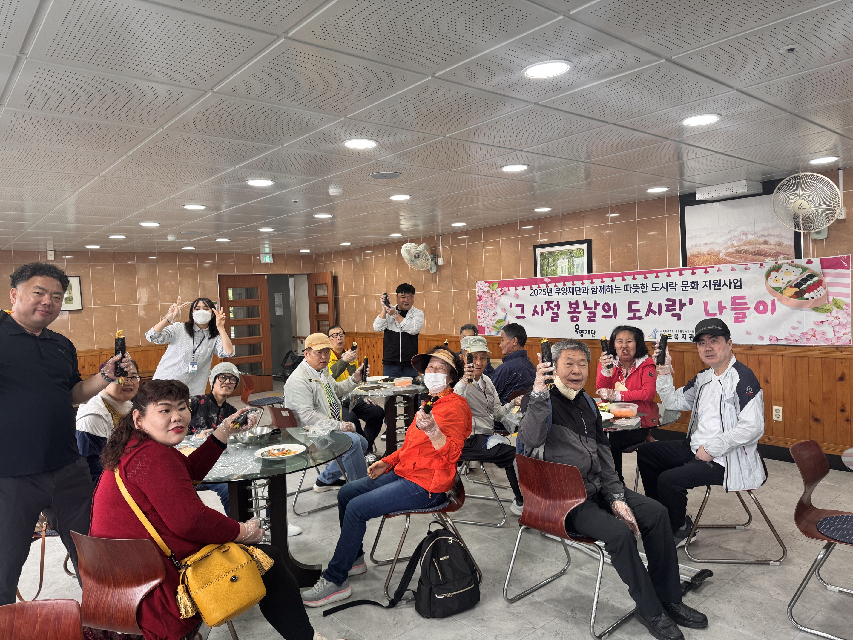 2025년 우양재단과 함께하는 따뜻한 도시락 문화지원사업. 그시절 봄날의 도시락 나들이라고 적힌 현수막이 관내 식당 벽면에 붙여 있고,  그 앞으로 김밥만들기를 함께한 재가장애인분들과 직원들이 모두 함께 자리에 앉아 완성된 김밥 한줄씩을 들고 찍은 단체사진입니다. 