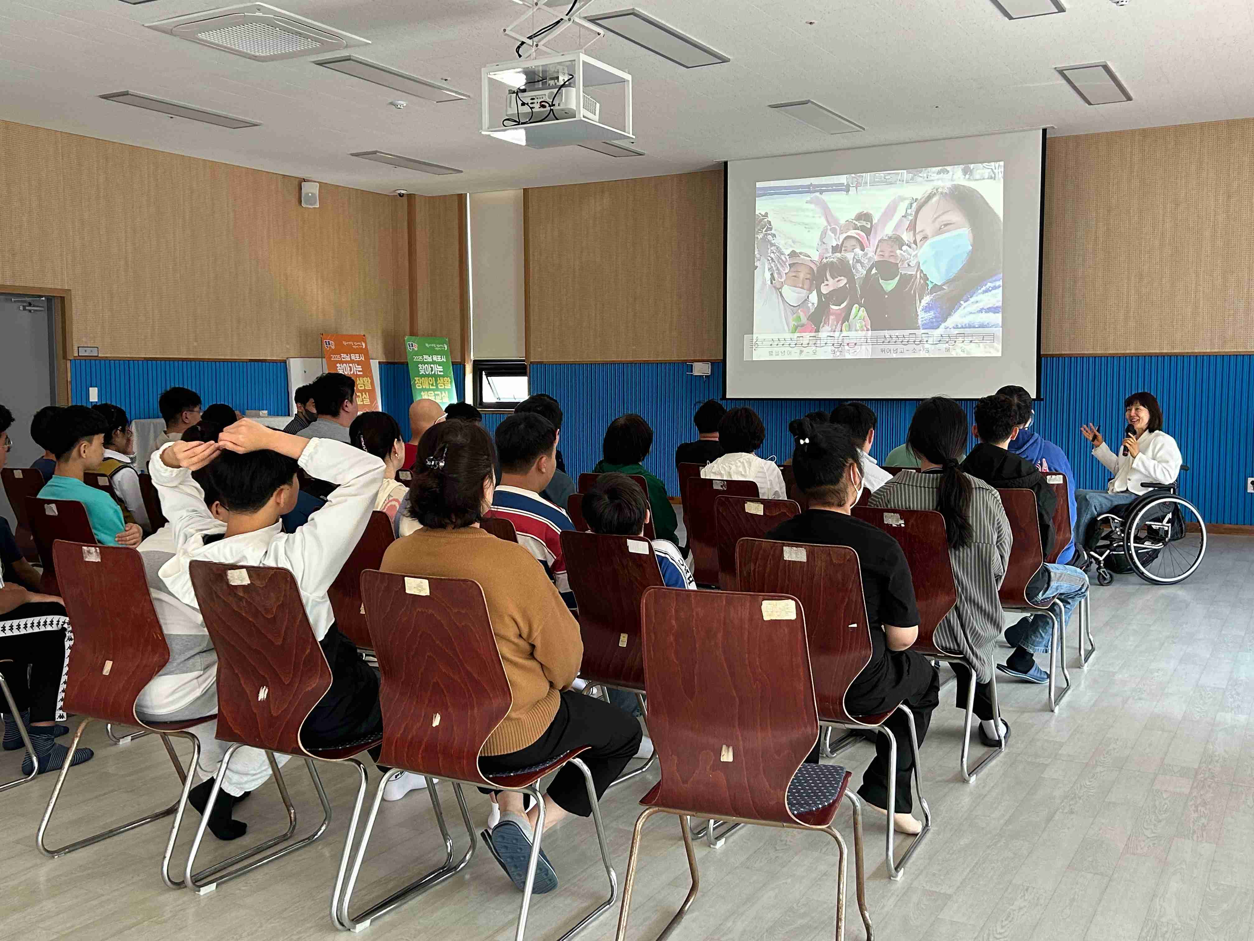 4동 강당에서 많은 이용인들이 의자에 앉아 강의를 듣고 있는 뒷모습입니다.