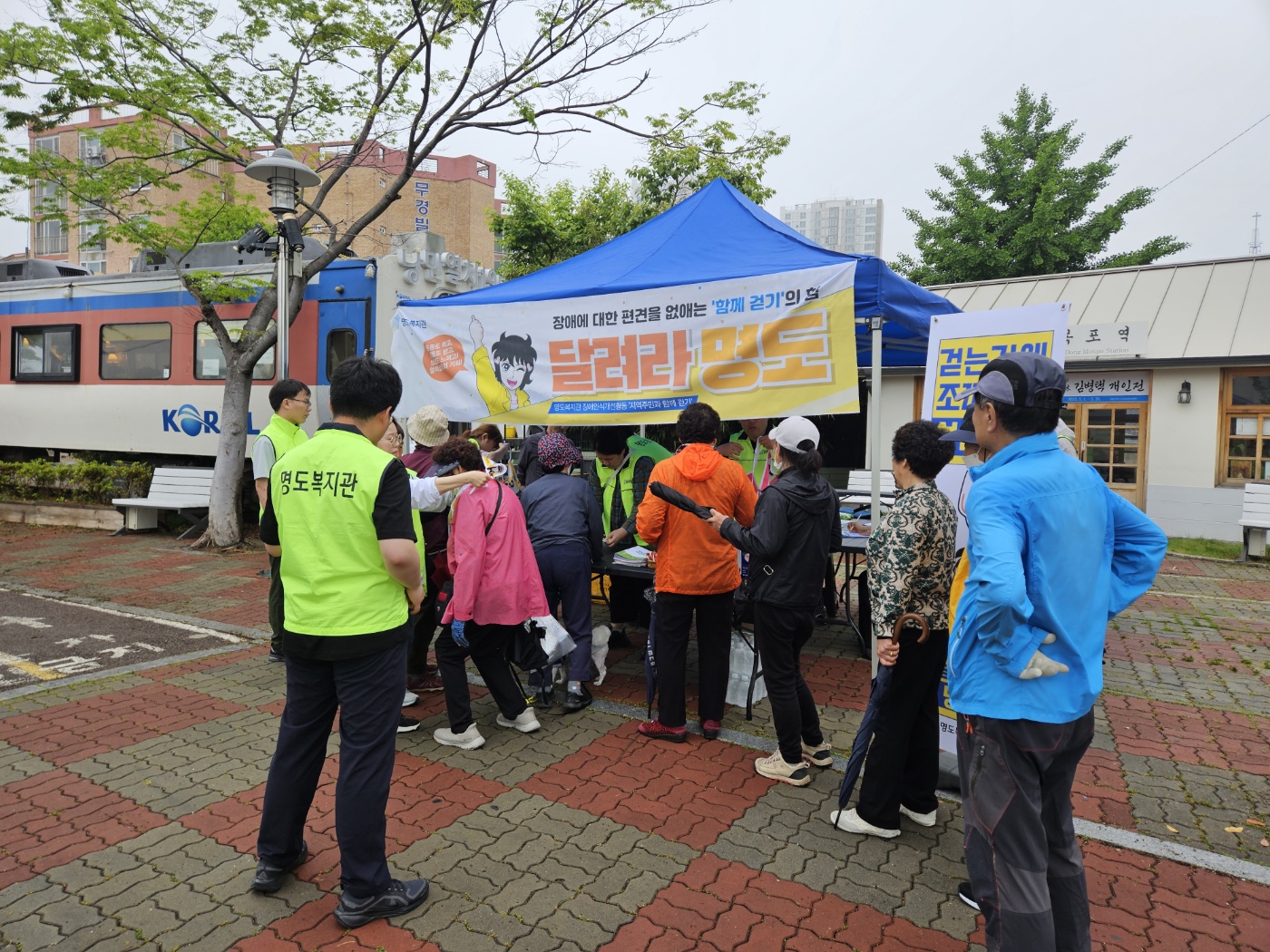 '달려라명도'라고 적힌 현수막이 걸린 천막아래에서 지역주민들이 접수를 하고 있는 모습의 사진입니다.