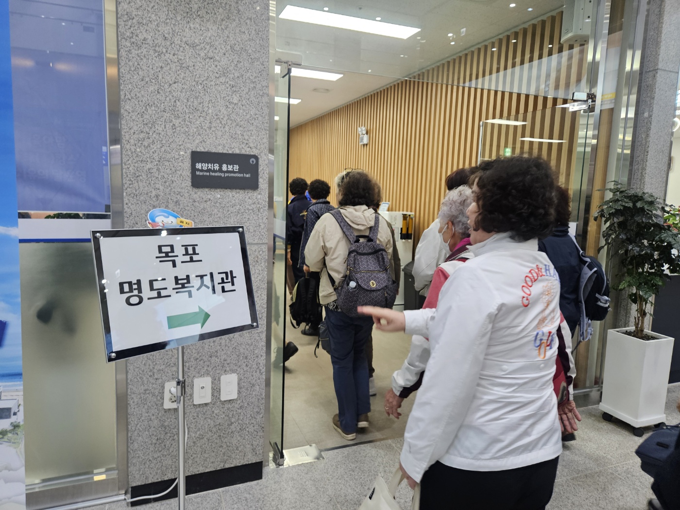 완도 해양치유센터 내 해양치유 홍보관에 입장하고 있는 자원봉사자들의 모습입니다. 한켠에는 자원봉사자를 안내하기 위해 명도복지과 화살표가 삽입된 안내판이 놓여져 있습니다.