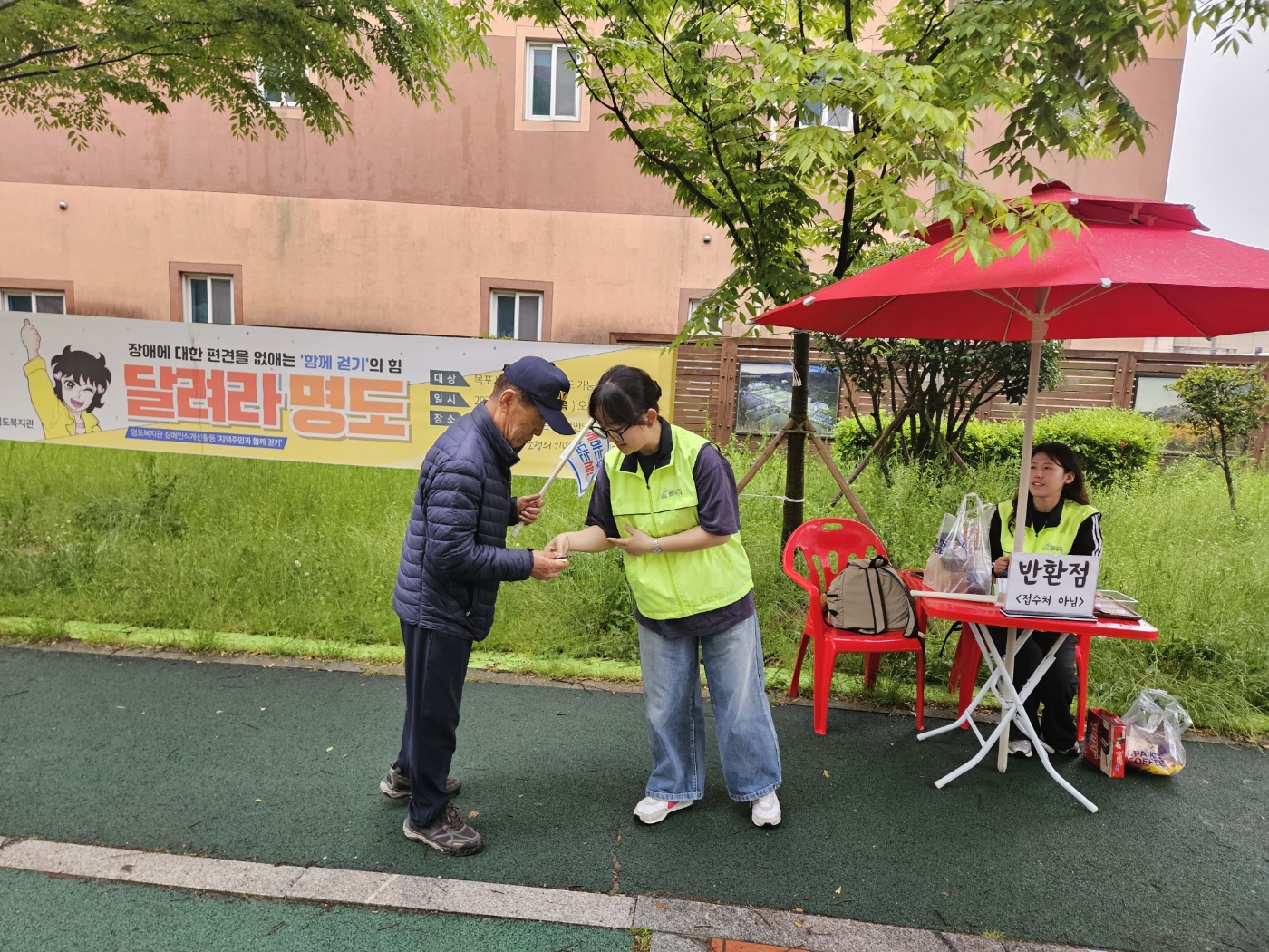 달려라명도 현수막 옆에 설치된 반환점에서 간식을 주고받고 있는 지역주민과 자원봉사자 모습의 사진입니다. 한명의 자원봉사자는 자리에 앉아 있습니다.