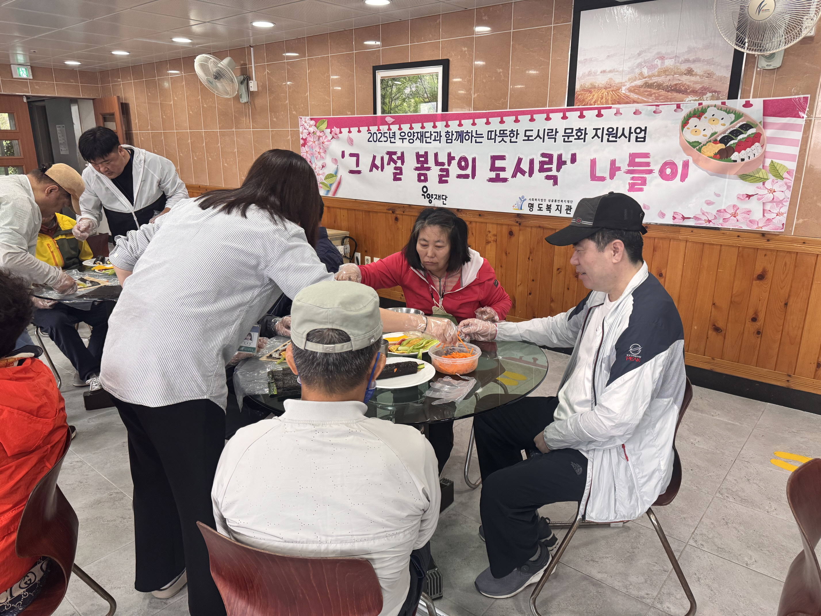 2025년 우양재단과 함께하는 따뜻한 도시락 문화지원사업. 그시절 봄날의 도시락 나들이라고 적힌 현수막 앞에서 재가장애인분들과 담당 사회복지사가 김밥을 싸고 있는 모습입니다. 