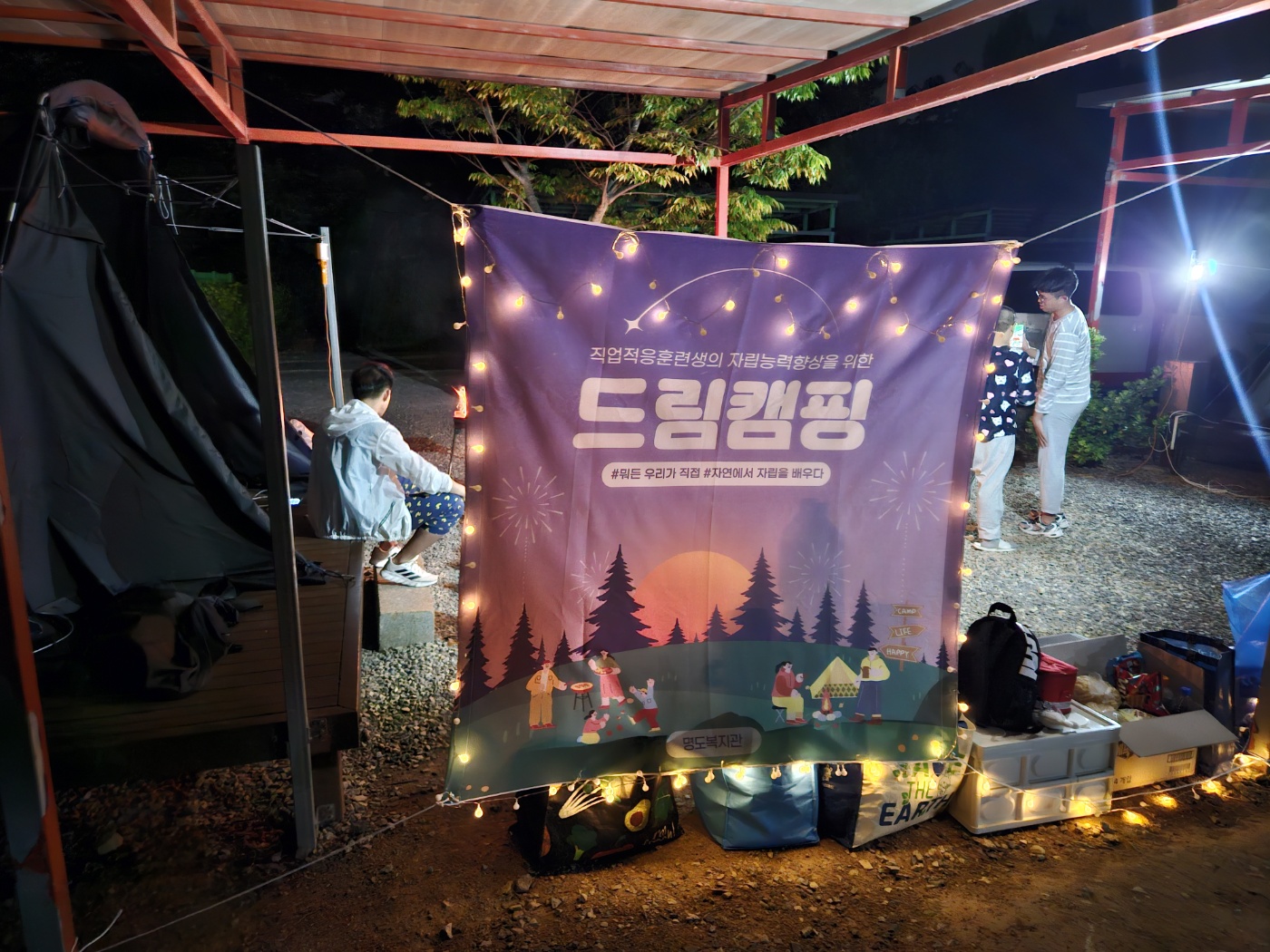 배경은 밤이고, 텐트뒤로 걸린 드림캠핑 현수막 위로 감성조명을 달아 예쁘게 반짝이고 있는 사진입니다. 