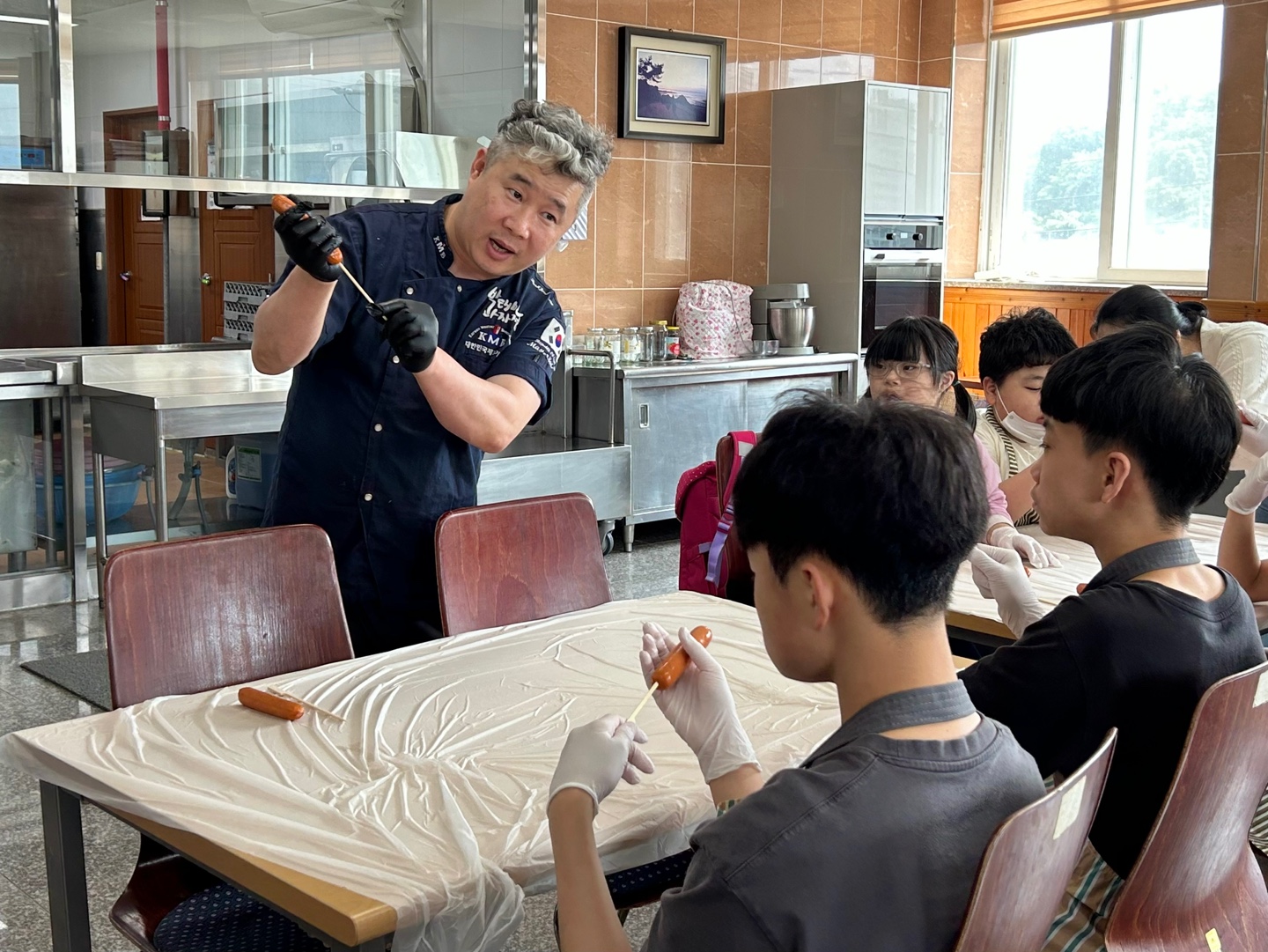 기능장님께서 소세지를 나무젓가락에 꽂는 시연을 하고 있고 방과후교실 아이들이 집중해서 따라하고 있는 모습입니다. 