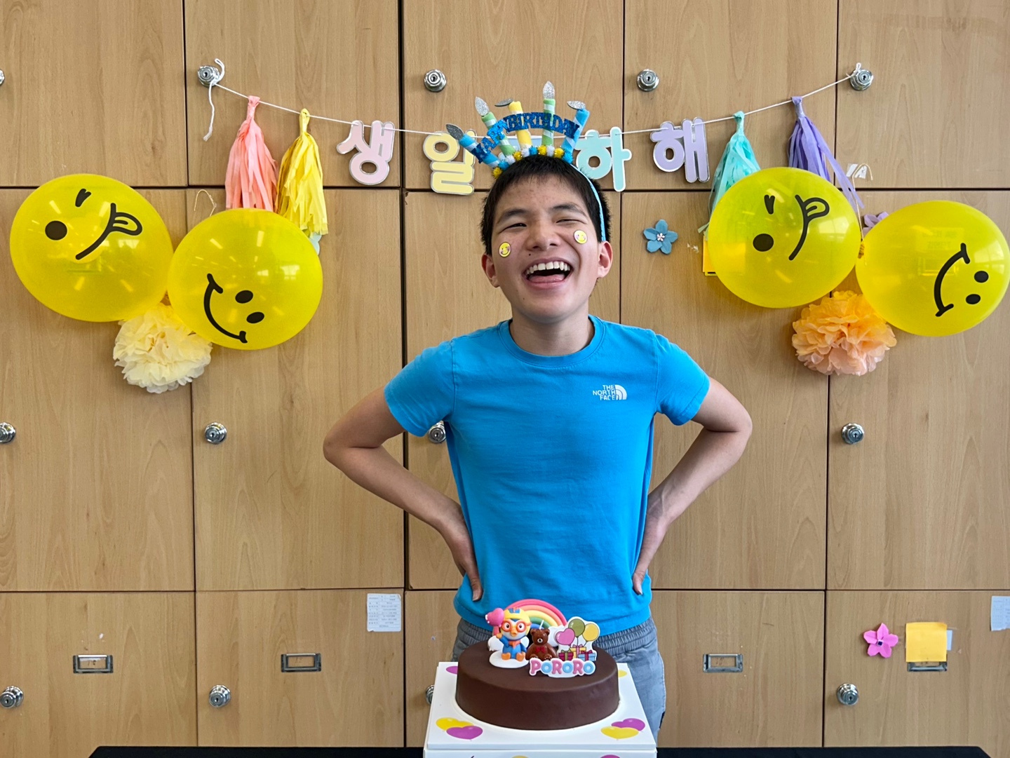 생일축하해라는 가랜드와 노란 풍선으로 장식된 벽을 배경으로 케이크를 앞에 두고 밝게 웃고 있는 이용인의 모습입니다. 