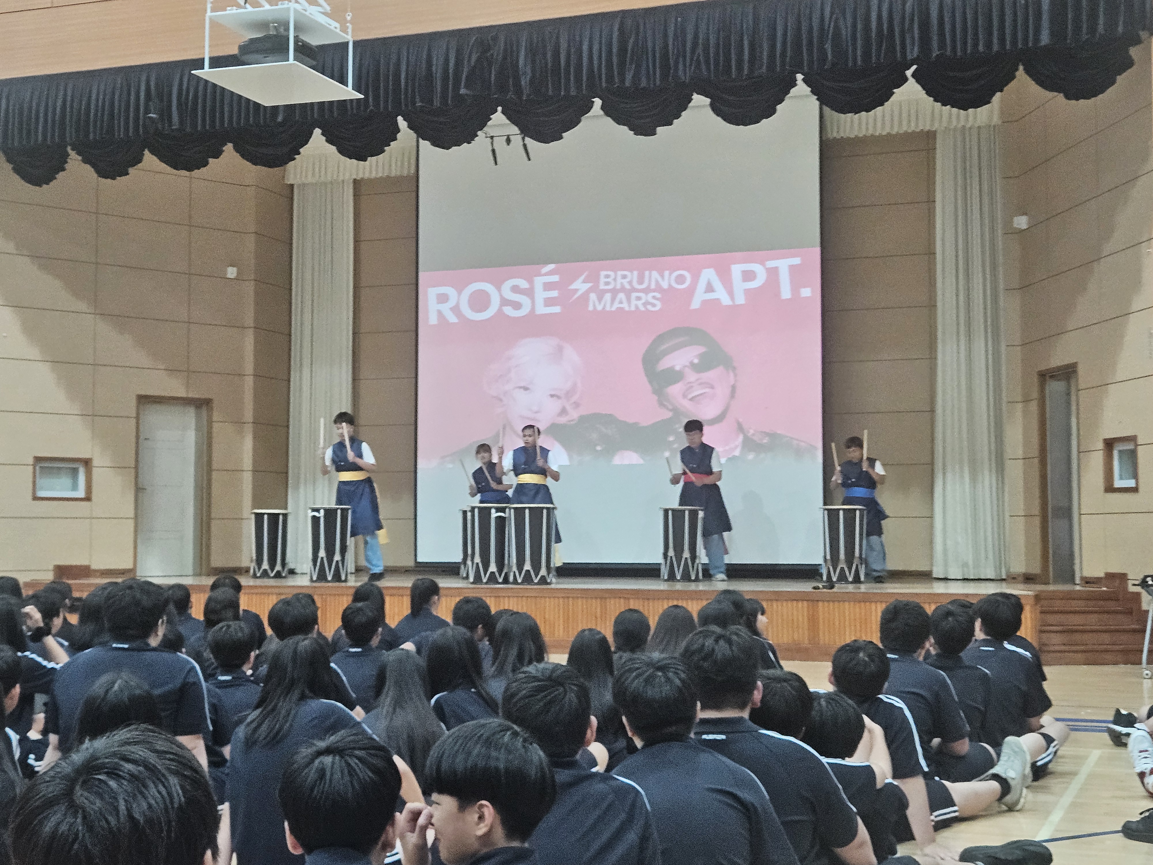 강당의 무대에서는 리듬공감팀의 공연이 진행되고 있고, 아래에는 학생들이 집중해서 공연을 관람하고 있는 뒷모습의 사진입니다. 