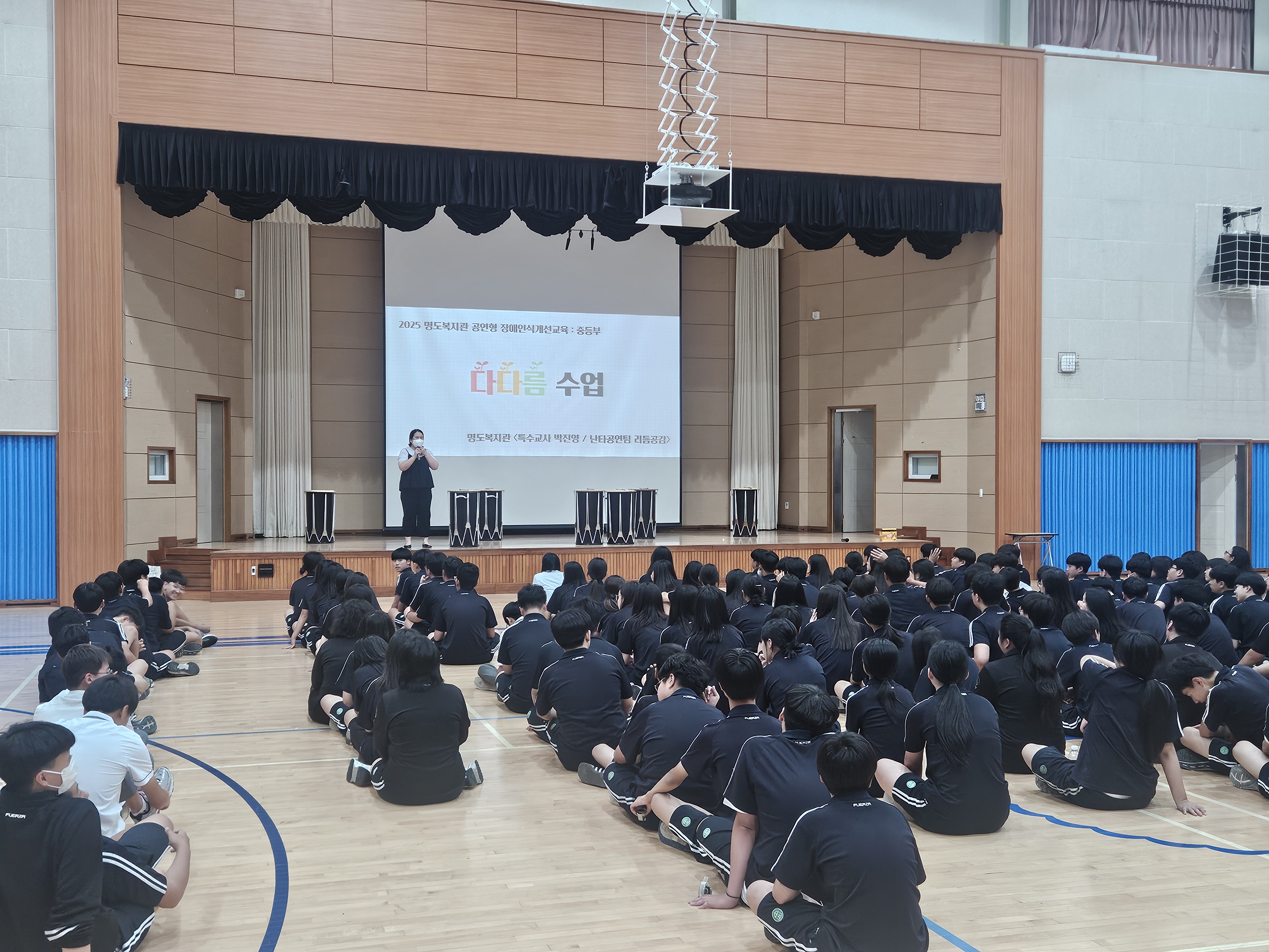 문태중학교 체육관에 학생들이 앉아 있고, 명도복지관 장애인식개선 교육인 '다다름 교육'이 진행되고 있는 사진입니다. 