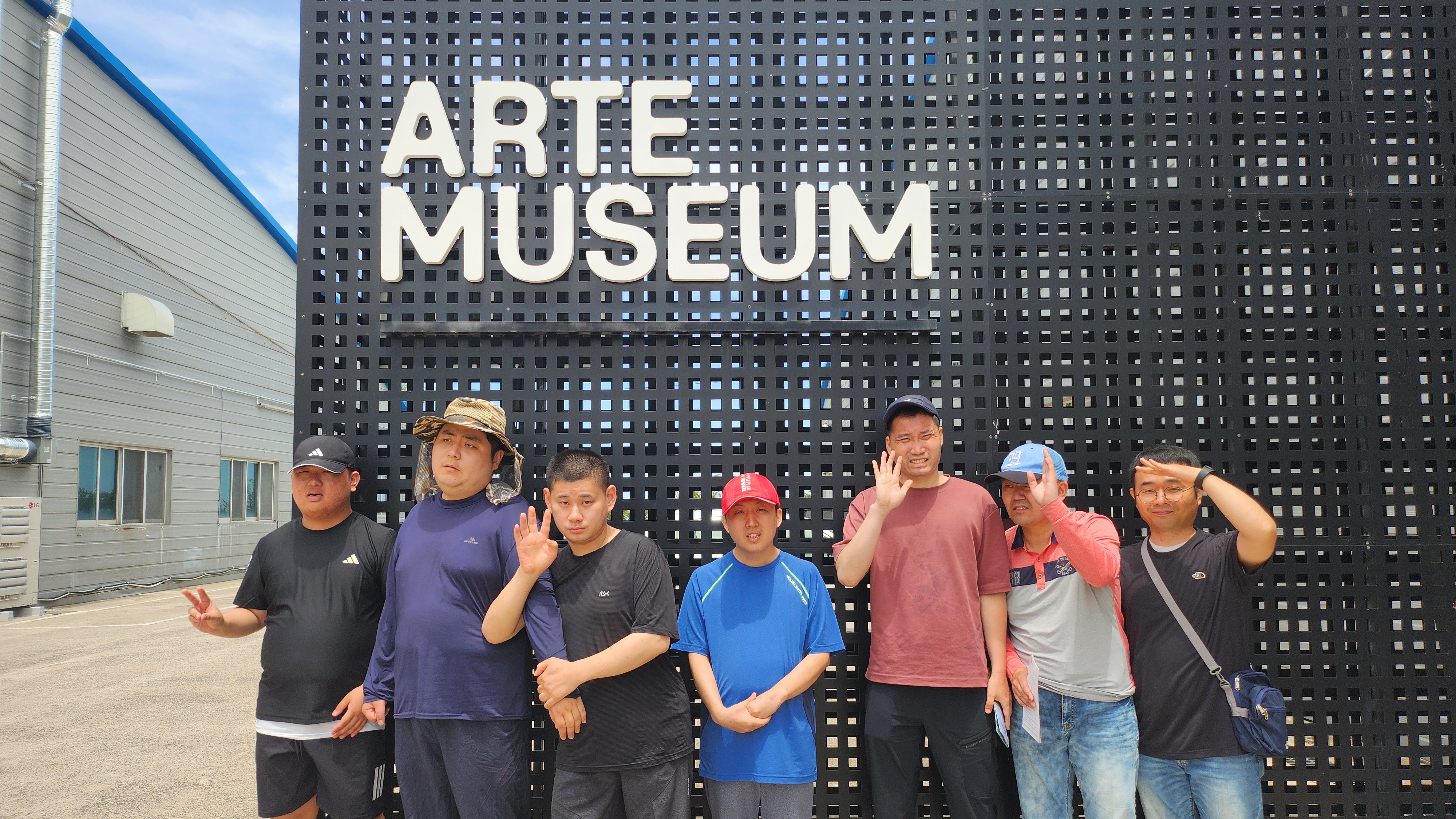 ARTE MUSEUM 이라는 글씨가 적혀있고, 그곳을 배경으로 주간보호센터 이용인과 교사가 브이 및 인사를 하며 단체사진을 찍고 있는 모습입니다.