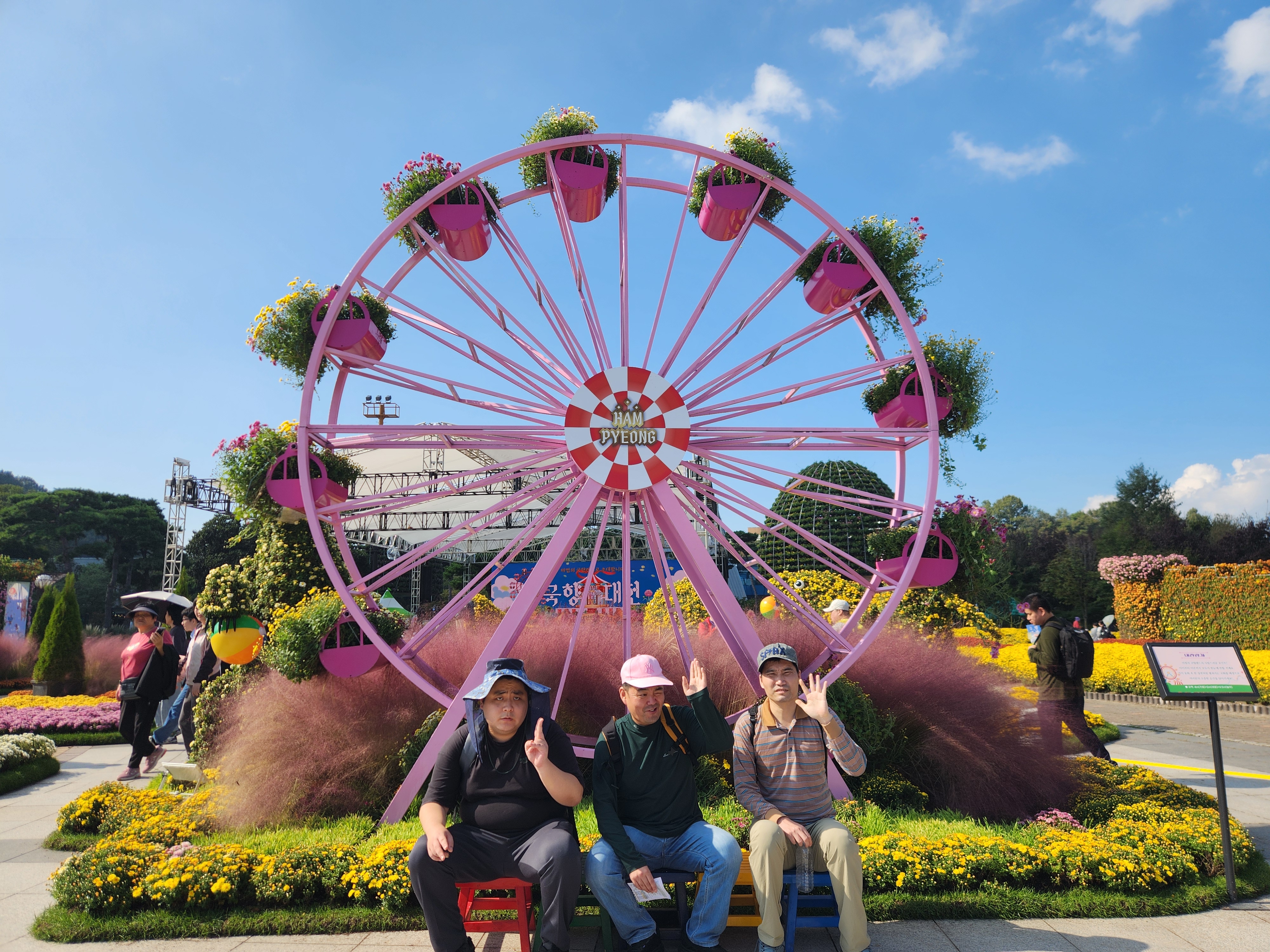남성 이용인 3명이 관람차 모양의 조형물 앞 의자에 앉아 각자 포즈를 취하고 있는 모습입니다.