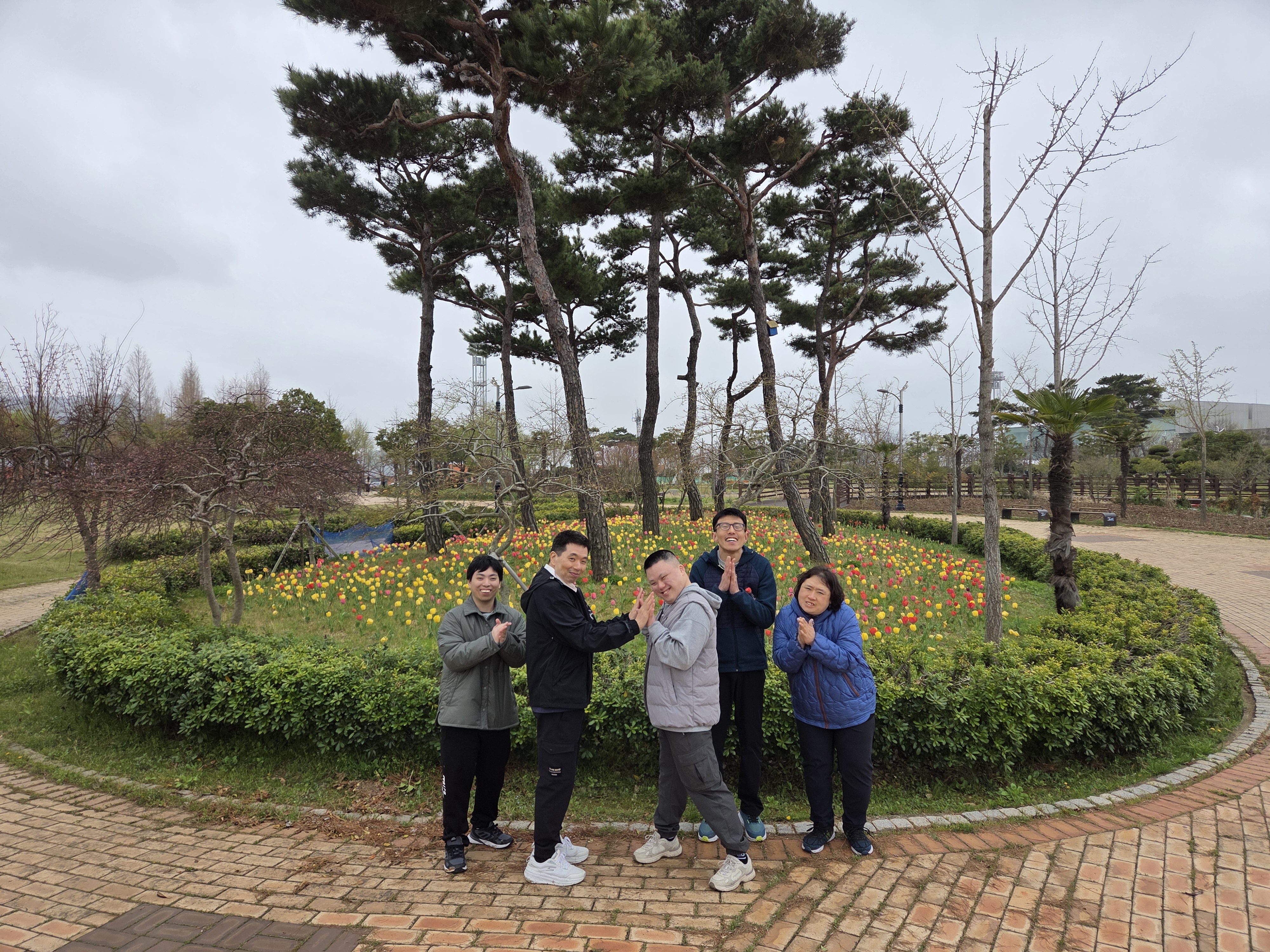 꽃밭 앞에서 2명의 이용인이 손을 맞대고 있고, 3명의 이용인은 박수를 치고있는 모습입니다.