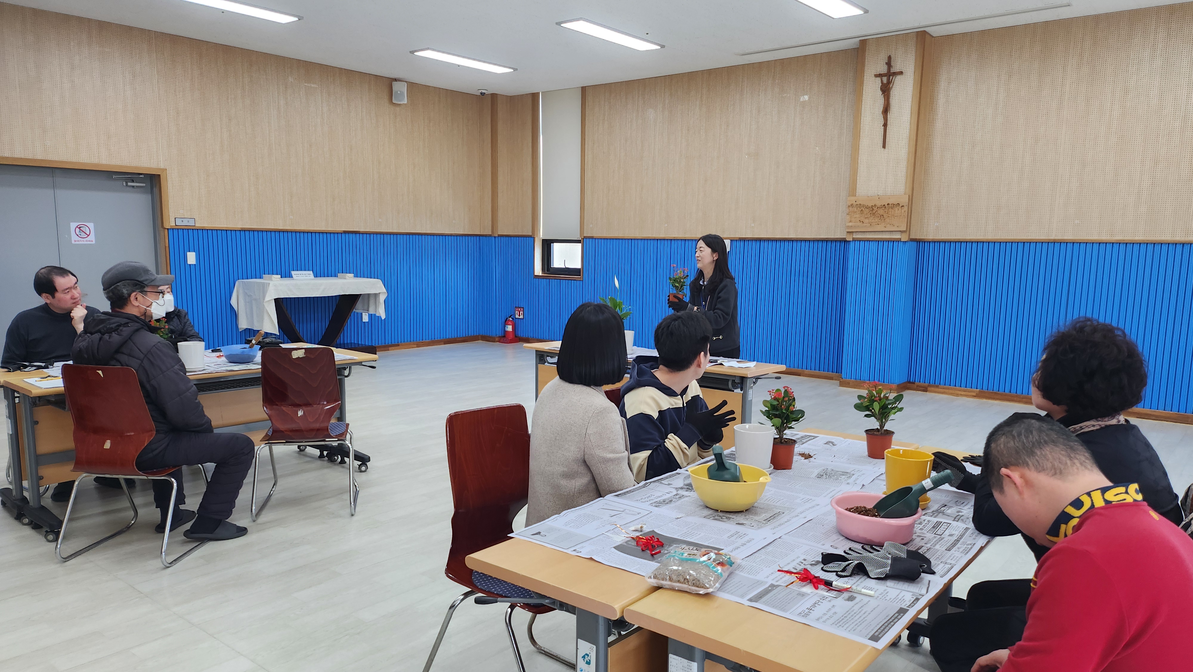 주간보호센터 선생님이 앞에 나와 화분을 들고 진행 방식에 대하여 설명하고 있고, 이용인분들과 보호자분들이 앉아 설명을 듣고 있는 모습입니다.